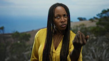 Sahil kenarında sarı gömlekli bir kadın açık havada okyanus arka planına karşı yoğun bir ifadeyle işaret ediyor.