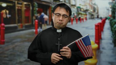 Amerikan bayrağını şehir caddesinde elinde tutan rahip kırmızı cüsseli ve bulanık şehir arkaplanıyla, çok kültürlü görünümü tasvir ediyor