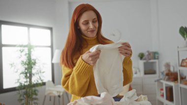 Evde, parlak bir çamaşırhanede elbiselerini katlayan bir kadın. Sarı bir kazak giyiyor. Gülümsüyor ve tatmin olduğunu gösteriyor..