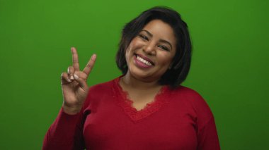 Kırmızı süveter giyen, yeşil arka planda barış işareti gösteren bir kadın pozitif ve canlı bir ortamda neşe ve güven ifade ediyor..