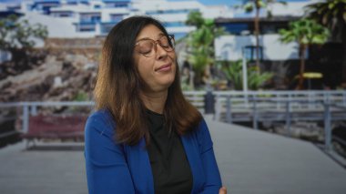 Gözlüklü ve kolları çaprazlanmış mavi ceketli bir kadın sokak geçidinde başını eğiyor..