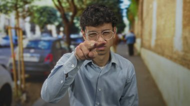 Gözlüklü genç İspanyol bir adam, şehir caddesinde park halindeki arabaların arasında kamerayı işaret ediyor..