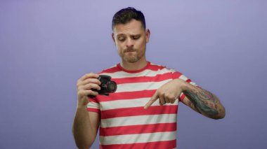 Dövmeli bir adam mor arka planda kamerayı tutuyor, aşağı bakıyor, çizgili gömlek giyiyor, kafa karışıklığı ya da merakını ifade ediyor..