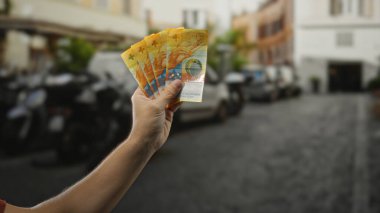Şehir sokaklarında İsviçre frangı tutan bir adam, banknotları şehir geçmişiyle belirgin bir şekilde sergiliyor, açık hava ortamında finansal bir temayı vurguluyor.