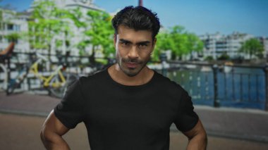 Adam şehrin ufuk çizgisinin altında silah akimbo ve sert ifade ile duruyor; kararlılık.