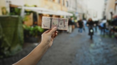 Meşgul bir şehir caddesinde Suudi Arabistan riyal banknotunu tutan bir adamın eli, canlı bir açık hava ortamının ortasında şehir hayatının ve ticaretin özünü yakalıyor..