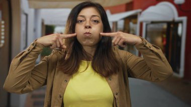Sarı bluzlu ve kahverengi ceketli kadın gün boyunca dudaklarını büzüştürürken parmaklarını yanaklarına işaret ediyor..