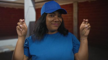 Kadın bitmemiş tuğladan bir binanın içinde mavi başlık ve gömlek giyerek el hareketi gösteriyor; şakacı ironi.