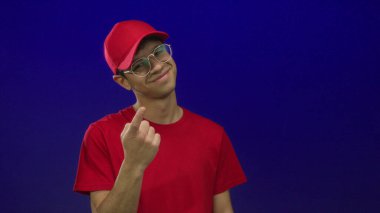 Gözlüklü, kırmızı şapkalı, gülümseyen ve mavi stüdyoyu işaret eden İspanyol genç adam..
