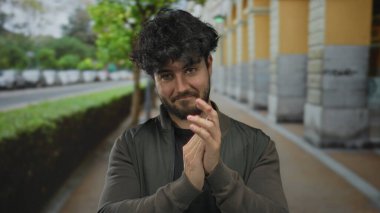Arka planda ağaçlar ve arabalarla şehir sokağında el çırpan sakallı genç İspanyol bir adam sıradan moda ve doğal ifade sergiliyor..