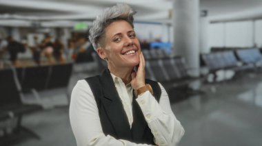 Yoğun havaalanı terminalinde siyah yelek ve beyaz bluzlu bir kadın kollarını kavuşturuyor..