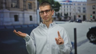 Gözlüklü bir adam sokakta parmak sallıyor. Açık hava ortamında tasvip etmediğini söylüyor. Arka planda binaları olan çizgili bir gömlek giyiyor..