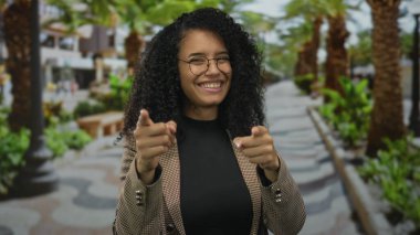 Genç İspanyol kadın, palmiye ağaçlarıyla kaplı güneşli bir parkta kamerayı işaret ediyor; güven bağlantısı nişan daveti.