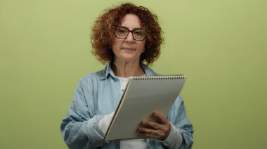 Orta yaşlı bir kadın not defterine izole edilmiş sarı arka plana karşı yazıyor, gözlük takıyor ve günlük kot gömlek giyiyor..