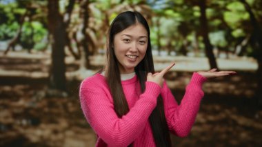 Pembe kazaklı genç bir kadın, güneşli bir park ortamında gülüyor, yemyeşil ağaçlarla çevrili, neşeli ve rahat bir açık hava sergiliyor..