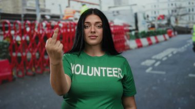 Yeşil tişörtlü kadın şehir sokaklarının arka planında duruyor. İnşaat bariyerleri ve sokak sahneleri arasında el hareketleriyle meydan okuyor.