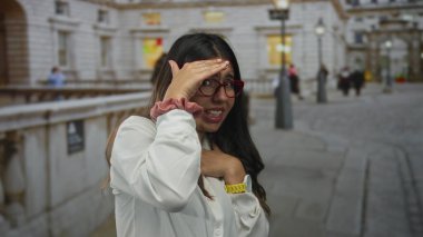 Gözlük takan ve sandık taşıyan bir kadın şehir dışında gündüz şehir ortamında arka planda ve tarihi mimaride insanlarla duygu gösterisi yapıyor..