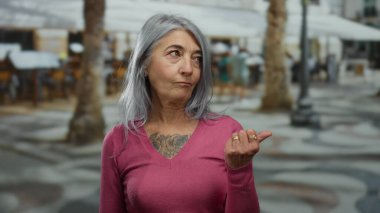 Gri saçlı ve pembe kazaklı bir kadın arka planda bulanık bir şekilde oturuyor ve ağaçlar baş parmağıyla yan tarafları işaret ediyor..