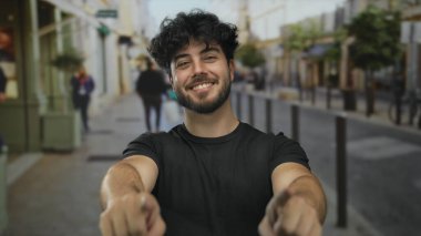 Sakallı genç İspanyol adam güvenli bir şekilde dışarıda poz veriyor. Canlı caddede kollarını kavuşturuyor ve kameraya gülümsüyor. Pozitif tutum ve kentsel yaşam tarzını gösteriyor.