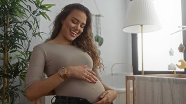 Bebek mobilyası ve bitkilerle çevrili rahat bir yatak odasında karnına hafifçe dokunarak gülümseyen hamile kadın annelik için neşe ve beklenti saçıyor..