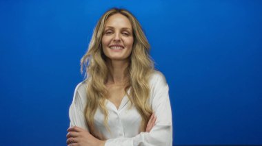 Kolları parlak mavi bir arkaplanda durarak poz veren kadın, beyaz bir gömlek giyiyor ve sakin bir ifade sergiliyor, modern sadeliği ve zarafeti vurguluyor..