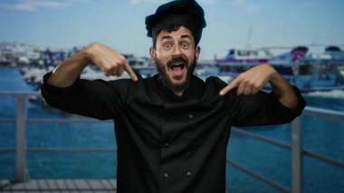 Siyah üniformalı bir şef gibi giyinmiş İspanyol sakallı adam arka planda gemiler ve teknelerle dolu bir limanın yakınlarında neşeli bir şekilde el kol hareketi yapıyor..