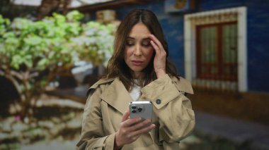 Şehir caddesinde akıllı telefon kullanan bir kadın arka planda bir ağaçla stresli bir şekilde ortaya çıkıyor ve elini şehir ortamında açık havada yüzünü kapatırken duygularını sergiliyor..