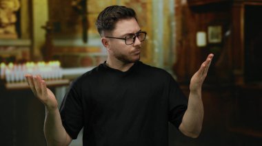Sakallı ve gözlüklü genç bir adam kilise ortamında dikiliyor, kararsız bir şekilde arka planda bulanık mumlarla loş bir çevrede avuçlarına bakıyor..
