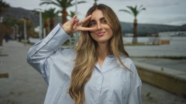 Sarışın genç bir kadın, palmiye ağaçlarıyla dolu bir marina güvertesinde, parmaklarıyla bir barış işareti tutuyor..