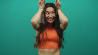 Turuncu bluz giyen gülümseyen İspanyol kadın stüdyoda parmaklarıyla tavşan kulağı hareketi yapıyor..