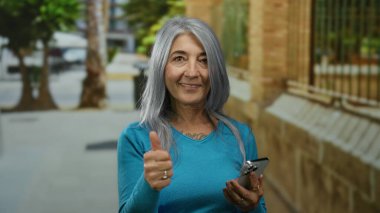 Gri saçlı, gülümseyen ve baş parmak kaldıran yaşlı bir kadın güneşli bir caddede akıllı bir telefon kullanırken teknolojiden anlayan neşe ve kendine güveni sergiliyor..