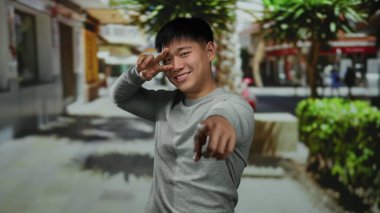 Açık şehir sokak ortamında gülümseyen genç Çinli bir adam hareketli bir şehir ortamında canlı bir yeşilliğin altında canlı bir ifade sergiliyor..