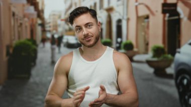 Şehir sokağında sakallı genç bir adam. Üzerinde beyaz bir atlet var. Şehrin arka planında binalar ve arabalar var..