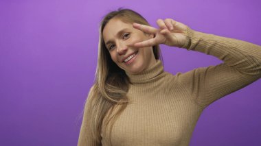 Mor arka planda barış işareti yapan, kahverengi balıkçı yaka giyen bir kadın, dışa dönük görsel kavramlar için ideal ortamda neşeli, pozitif bir hava yaratıyor.