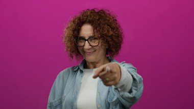 Canlı pembe bir arka plana gülümseyen, elleriyle kalp şekli oluşturan, gözlük takan, kot gömlek giyen, stüdyoda mutluluk ve pozitiflik gösteren bir kadın..