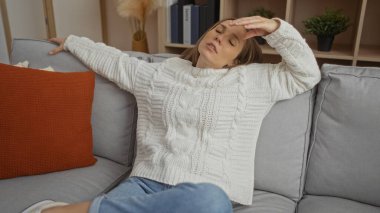 Rahat bir oturma odasında rahat bir kanepede dinlenen, beyaz bir kazak giyen, yorgunluk ifadesi olan, rahat dekorlar ve yumuşak yastıklarla çevrili genç bir kadın..