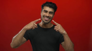 Siyah tişört giyen genç İspanyol adam kırmızı stüdyo ışıkları altında geniş bir gülümsemeyi işaret ediyor..