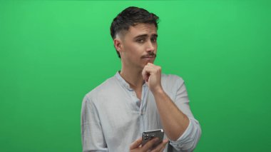 Genç adam canlı yeşil arka plan önünde akıllı telefonu tutarken, modern teknolojiyi ve içsel düşünceleri tasvir ederken, gündelik açık gömlekler giyerken,.