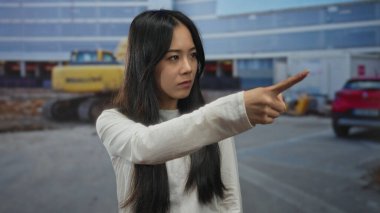 Kadın gün ışığında inşaat alanını kapalı gözlerle işaret ediyor, beyaz gömlek giyiyor, sanayi ortamında yön veya karar verme mekanizmasını temsil ediyor..