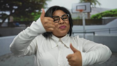 Genç, iri yarı bir kadın basketbol sahasında başparmağını kaldırıyor, kendine güvenerek gülümsüyor, gözlük ve beyaz gömlek giyiyor, pozitiflik ve kendine güvenini gösteriyor..