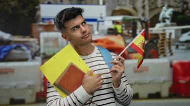 İspanyol bayrağı ve klasörleri taşıyan adam, şehrin bir caddesindeki inşaat sahasında düşünceli bir şekilde duruyor..