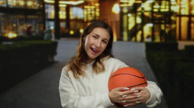 Sokakta elinde basketbol topuyla gülümseyen genç bir kadın aydınlanmış arka planda, açık hava kentsel ortamının tadını çıkarıyor..