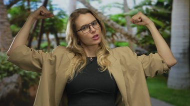 Gözlüklü, kendine güvenen sarışın bir kadın yeşil bir parkta kaslarını esnetiyor. Günlük bir spor ceket giyiyor. Canlı, doğal bir ortamda güç ve güç yayıyor..