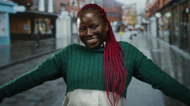 Yağmurlu bir şehir caddesinde kollarını açmış gülümseyen bir kadın şehir ortamında neşesini ve varlığını vurguluyor..