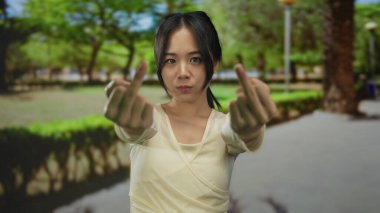 Parktaki genç Çinli kadın, güneşli bir günde çevresini çevreleyen canlı yeşil ağaçlar ve açık havada kendinden emin bir ifadeyle orta parmak hareketi gösteriyor..