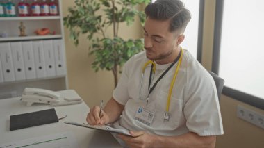 Sakallı genç bir doktor, parlak bir klinik odasında notlar yazıyor. Steteskop ve beyaz önlük giyiyor. Evrak işleri ve organizasyonla dolu profesyonel bir tıbbi ortama işaret ediyor..