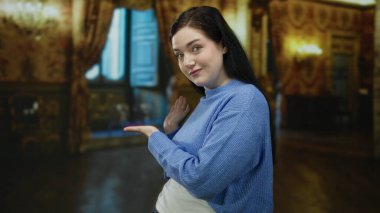 Mavi kazaklı kadın zarif bir sanat galerisinde süslü dekorlu ve dramatik ışıklandırmalı bir şekilde poz vererek büyüleyici ifadesini ve şık arkaplanını vurguluyor..