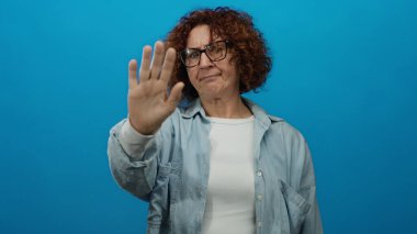 Kot pantolonlu kadın, ciddi ifadesini ve günlük kıyafetlerini vurgulayan canlı mavi bir duvara el kaldırıyor..