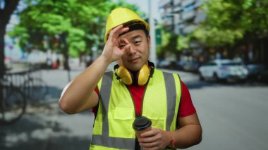 Güvenlik kaskı ve yelek giyen gülümseyen adam gündüz vakti elinde kahve tutuyordu.