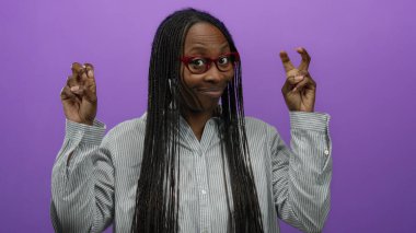 Mor duvara karşı kıvırcık saçlı ve gözlüklü el hareketleri yapan bir kadın neşeli bir ifade ve tavır sergiliyor..